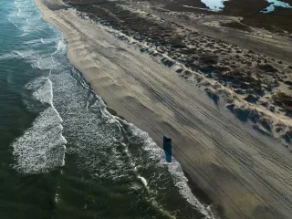 Drone shot of jeep driving on beach with ocean to the left.