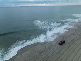 Drone shot of jeep driving on beach with ocean to the left in winter.