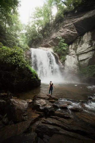 A person stands on rocks near the base of a large, misty waterfall surrounded by lush green trees and steep rocky cliffs.