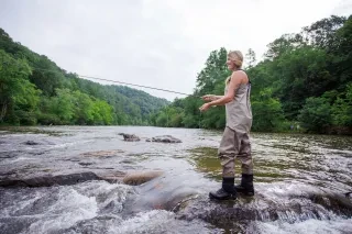 Woman fly-fishing in river surrounded by trees during daytime.