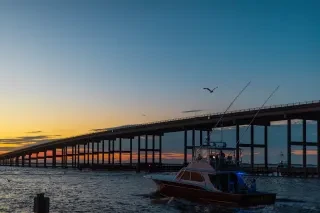 Fishing charter heading out into ocean alongside bridge during sunrise.