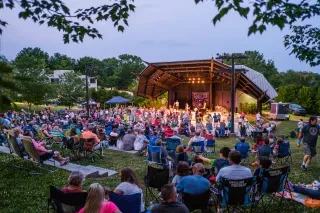 Crowd of people sitting on amphitheater lawn watching band perform at dusk.