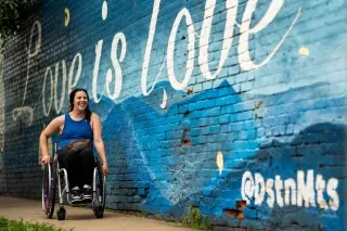 A smiling woman who is a wheelchair user moves along a sidewalk beside the "Love is Love" mural in the NoDa district of Charlotte, North Carolina.