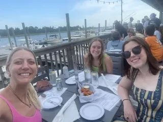 Three friends sitting at dockside restaurant taking a selfie with water and boats in background.