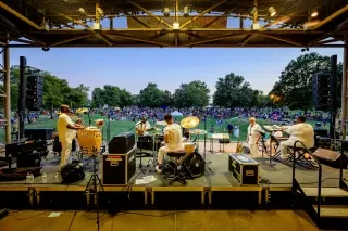 Band playing on stage looking out onto tons of people sitting on lawn enjoying show.