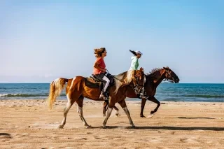Two women galloping on horseback on beach.