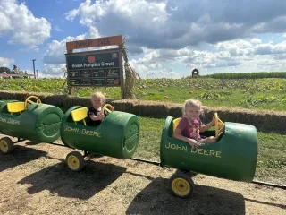 Two kids in tractor toys smiling at camera with pumpkin patch in background.