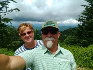 Husband and wife taking selfie on hiking trail with mountain views in background.