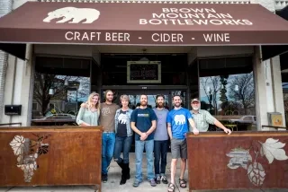 Friends posing for camera in front of Brown Mountain Bottleworks on outdoor patio under brown awning.