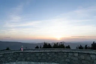 Woman walking on bridge within Mt. Mitchell State Park with sun setting over mountains in the distance.