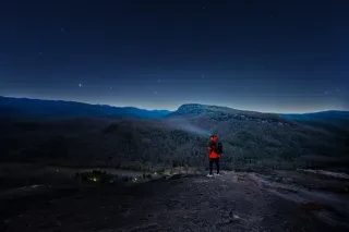 Person with light on head standing on mountain vista stargazing over mountains.