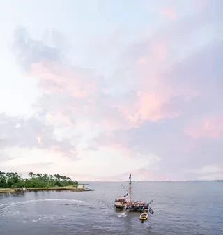 Aerial of pirate ship surrounded by calm waters under pink and blue skies.