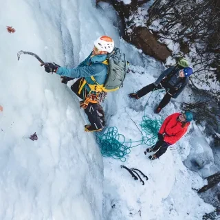 Man beginning to ice climb up wall of ice with people on ground watching him.