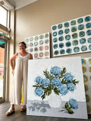 Artist Christie Younger stands next to large floral art with art on wall behind her.