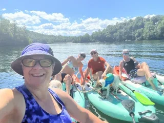 Woman taking selfie with three friends and a dog in kayaks on mountain lake.
