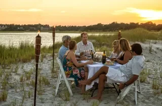 Friends dining at table on beach surrounded by torch lights, sand and sea oats at sunset.