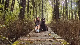 Girl giving peace sign and chocolate lab sitting on trail in forest surrounded by trees and shrubs.