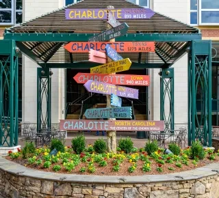 Fanciful sign denoting Charlotte, NC, as "the center of the known world." Flowers surround the sign, which is in front of a building.