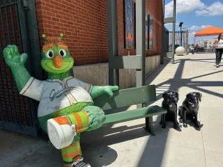 Guilford the Grasshopper mascot sitting on bench and waving at camera at baseball game.