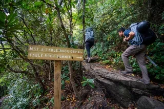 Two people carrying gear hiking up rock path with sign in foreground labeling trails.