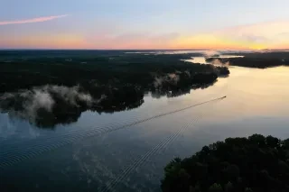 A fishing boat takes off into the sunrise on Hyco Lake. The sky is filled with bright pink and fades to blue. Fog rises from the lake.
