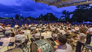 Symphony performing on stage in Fayetteville with large lawn of people in front of stage at dusk.