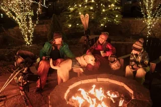 Family and dog sitting around ski resort fire at night surrounded by lit-up trees.