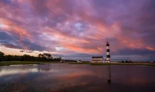 The Bodie Island Lighthouse stands on the right third of frame surrounded by a pink and purple sunset sky. It is reflected in shallow water at the base.