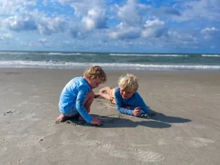 Two boys beachcombing in Onslow County under bright blue sky and ocean in background.