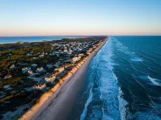 An aerial view shows oceanfront homes in Corolla lining the Outer Banks shoreline as sunlight illuminates the beach at sunrise.
