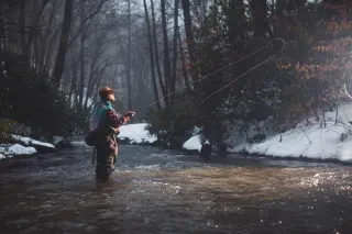 Man fly fishing in river surrounded by snowy land and bare trees in Boone during winter.