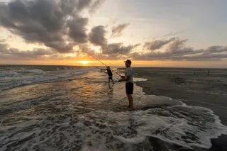 Two people stand in the surf fishing in the ocean as the sun sets behind them.