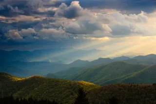 Blue Ridge mountains create layers of tree covered hills with the sun breaking through the clouds in the upper right corner.