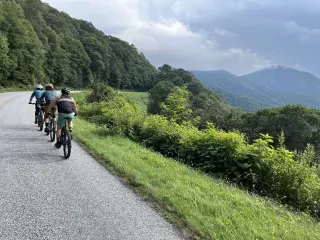 Three bikers on scenic mountain road with trees and mountain vistas to the right.