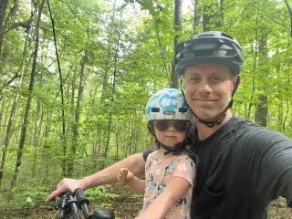 Man and toddler daughter wearing helmets and smiling for camera while biking on trail. 