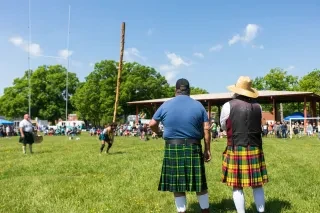 Men in kilts watch athlete competing in Highland Games event in field during daytime.