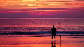 Silhouette of man surf-fishing on beach with bright pink and orange sunset.