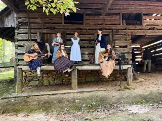 Women in period clothing singing and playing instruments in front of log cabin.