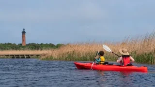 Woman and child in red kayak paddling through march with lighthouse in background.