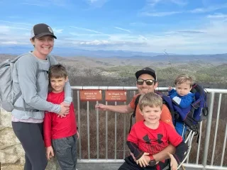 Family of 5 posing in front of blue mountains and green trees at overlook.