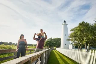 Family with child on dad's shoulders walking with Ocracoke Lighthouse in background.