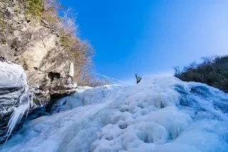 Man at top of frozen waterfall waving down to camera during daytime.