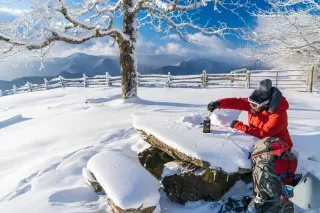 Man in winter gear sitting at snowy table with snow and mountains in background during daytime.