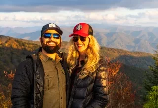 Couple wearing jackets and hats smiling at camera in front of scenic mountains during daytime.