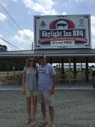 Man and woman smiling for camera in front of sign reading Skylight Inn BBQ.