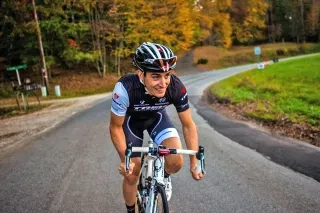 Man smiling while riding bicycle on road with fall foliage in background.