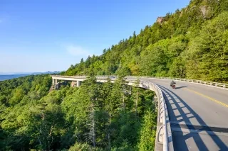 A motorcycle rides along a ling and winding road that hugs a mountain scape. 