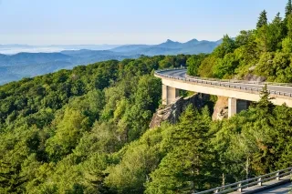 Linn Cove Viaduct to the right with green trees to the left and mountain ridges in background on clear day