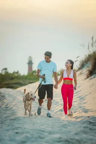 Couple walking dog on beach with lighthouse in background.